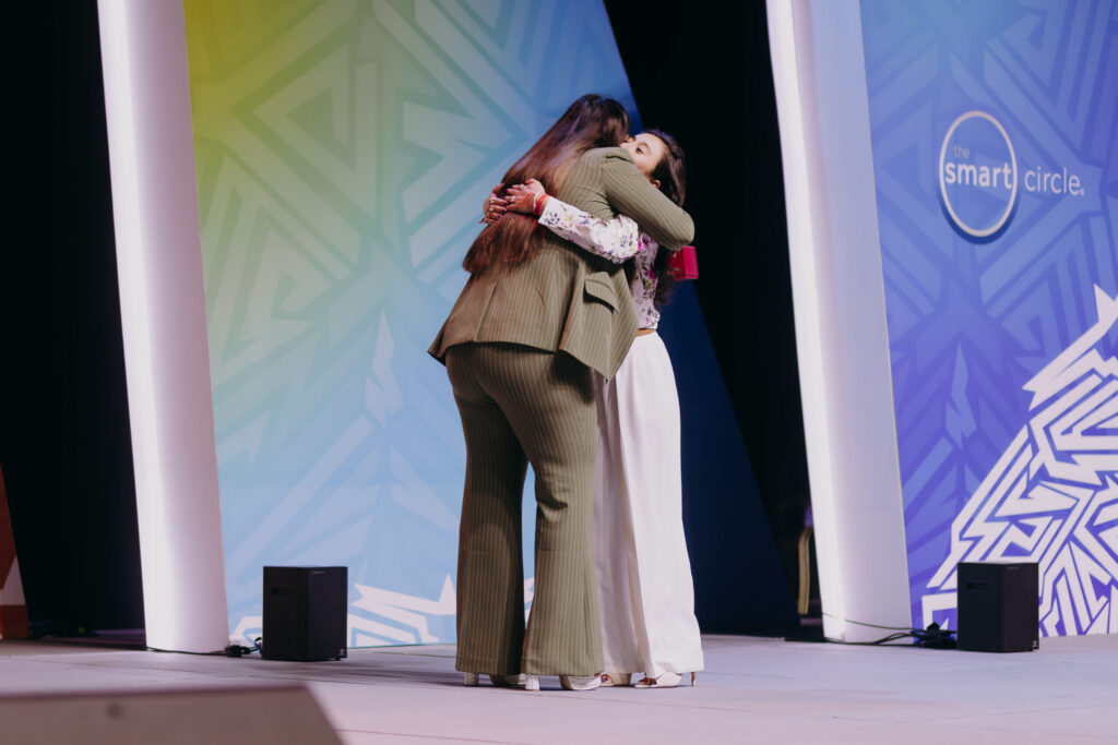 Two professional women hugging on stage during a Smart Circle corporate awards ceremony, representing company culture and excellence.